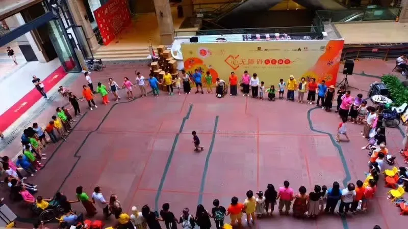 俯拍视角下,众多成人与儿童手拉手围成一个大圆圈,圈边有轮椅使用者,圆心空地上一人独自站立,背景是彩色的活动舞台背板。