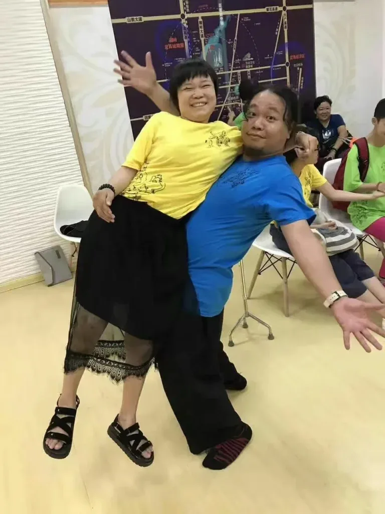 释文身穿黄色上衣,被丸仔搂在腰间抱起,两人张开手臂笑着摆出夸张姿势,身后有人坐在椅子上。