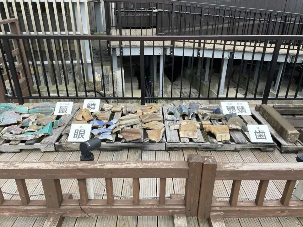木质展台上摆放着几组彩色琉璃瓦残片,残片旁立着写有“鳞”“眼”“顎”“歯”等汉字的白色标签牌,背景是施工脚手架护栏。