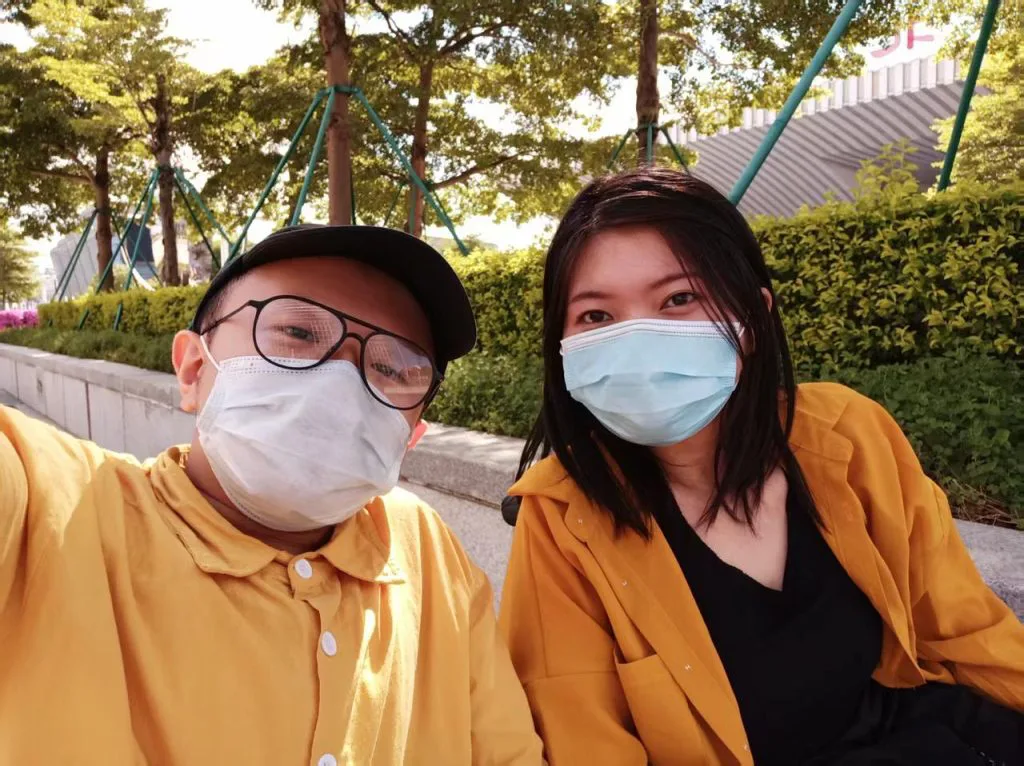 两人戴口罩并排坐在户外,一人戴鸭舌帽和眼镜,两人都穿黄色外套,身后是树丛草坪与攀爬架。