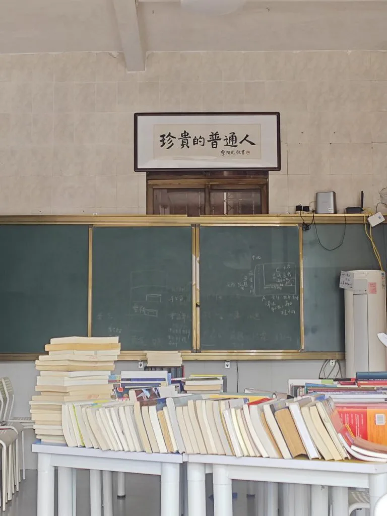 教室墙上挂着“珍贵的普通人”横幅,下方是绿色黑板,前景课桌上堆满书籍。