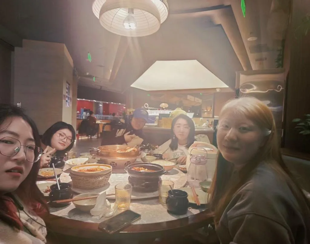 几位女性围坐圆桌用餐,桌上摆着火锅和多道菜肴,头顶吊灯洒下暖光。