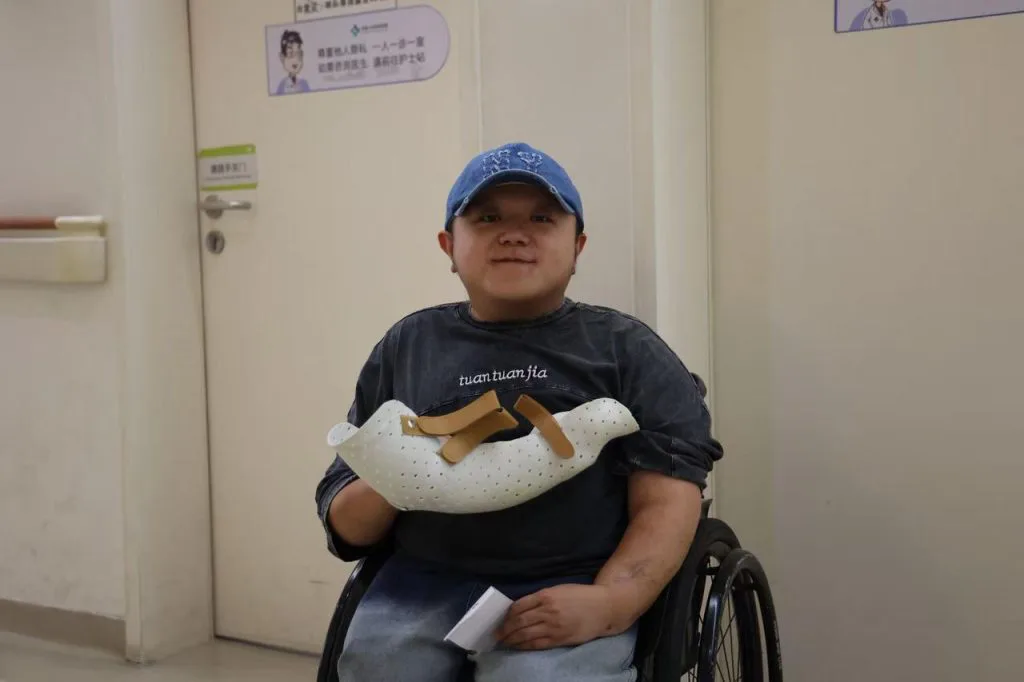 男子戴蓝色棒球帽坐在轮椅上微笑,手臂戴着白色镂空支具,身后是医院走廊。