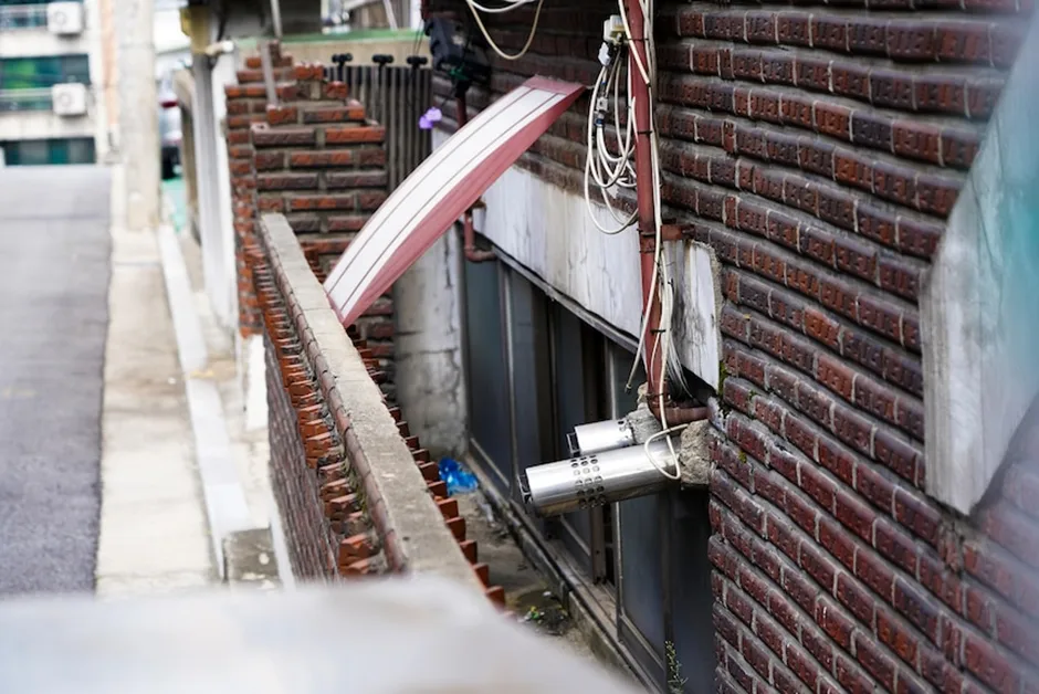 红砖建筑沿坡道而建，外墙缠绕着电线和管道，靠近地面处是带金属护栏的半地下室窗户，窗下堆着杂物。