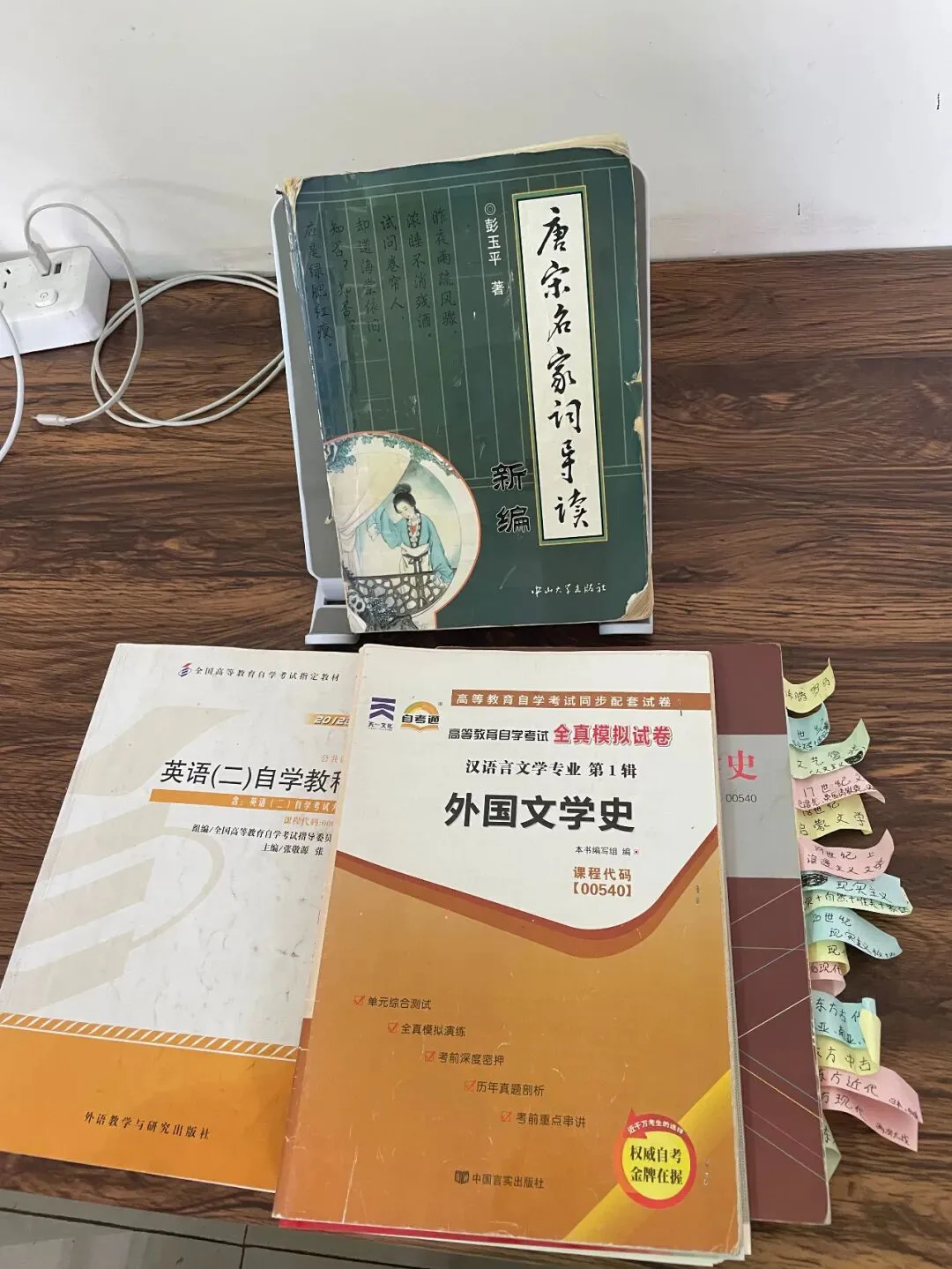 书桌上摆放自考备考书籍的照片，前排竖立《唐宋名家词导读新编》，其后是《英语（二）自学教程》和贴满彩色标签纸的《外国文学史》全真模拟试卷。