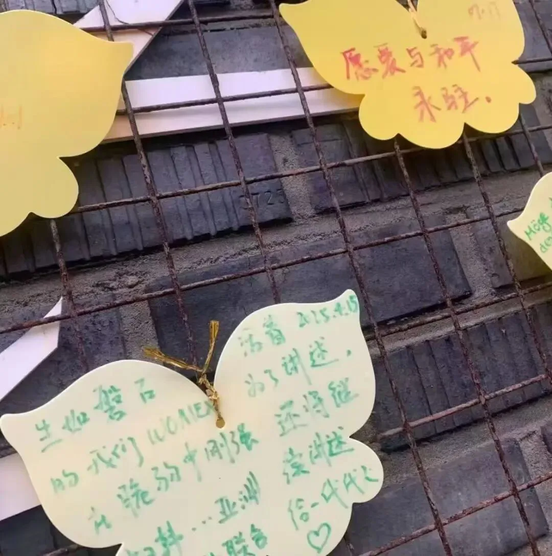 蝴蝶形彩色纸片系在锈迹斑斑的金属网格上，纸片上手写留言，其中一张写着“愿爱与和平永驻”，另一张提到“WOMEN”及持续讲述诉求，其余字迹潦草不易辨认。