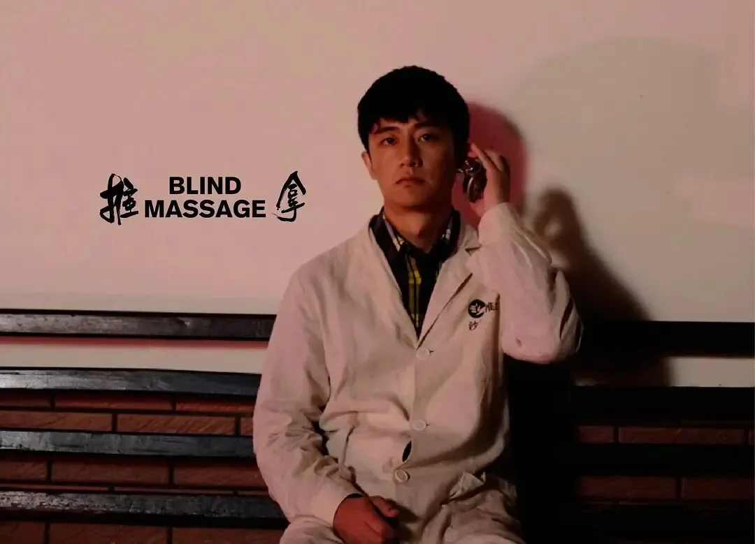 剧照中一名男子坐在木椅上，身穿米色外套，手持手机贴在耳边，目光望向前方；画面左侧印有片名《推拿》及英文 BLIND MASSAGE。