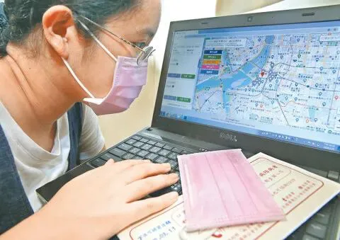 一位戴口罩的女性对着笔记本电脑查看地图页面,桌上放着一片粉色口罩和印有文字的购买凭证。