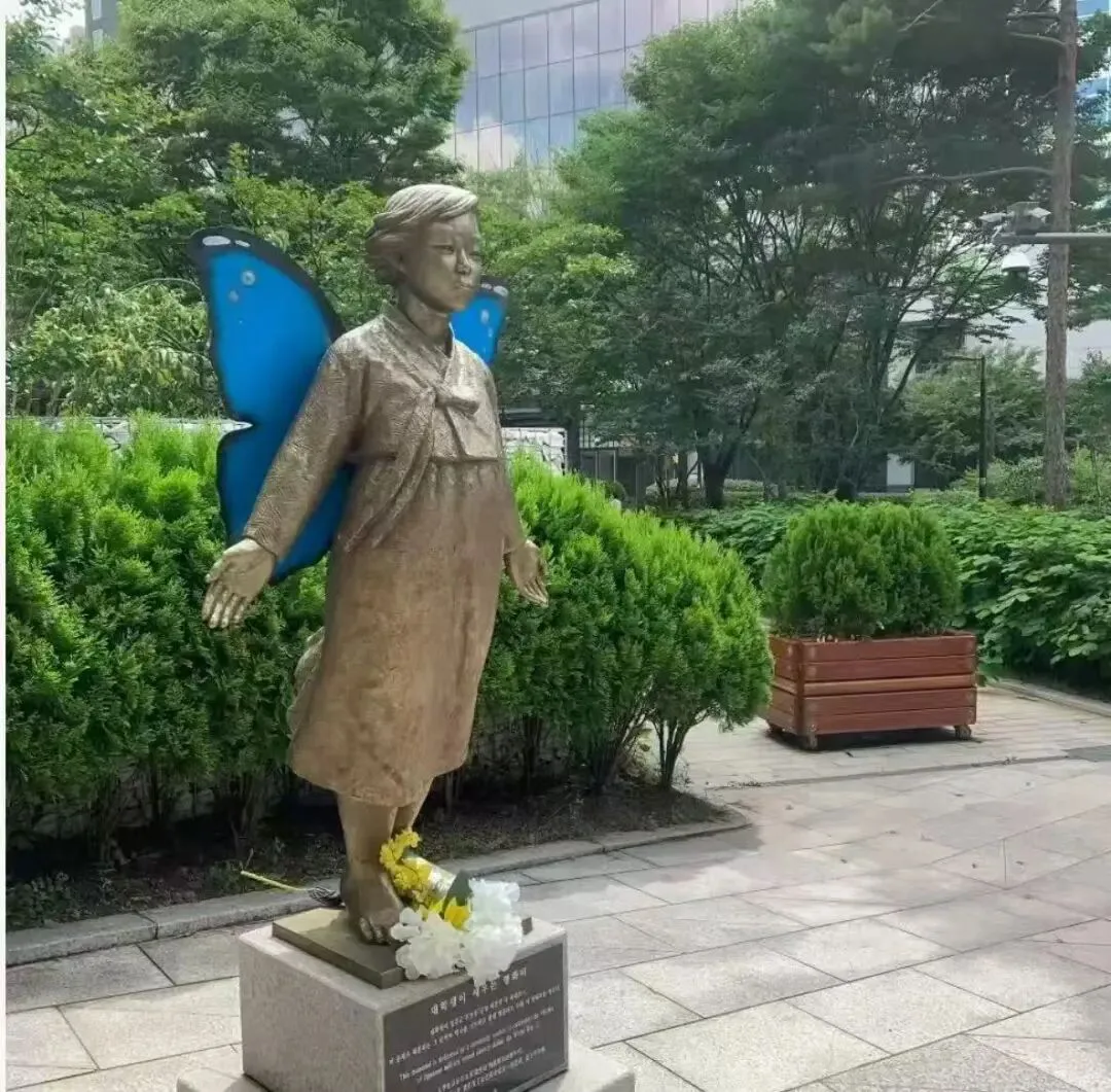 青铜色雕像身着传统韩服站立，身后展开一对蓝色蝴蝶翅膀，脚边摆放黄白色鲜花，四周环绕修剪整齐的绿植和石板广场。