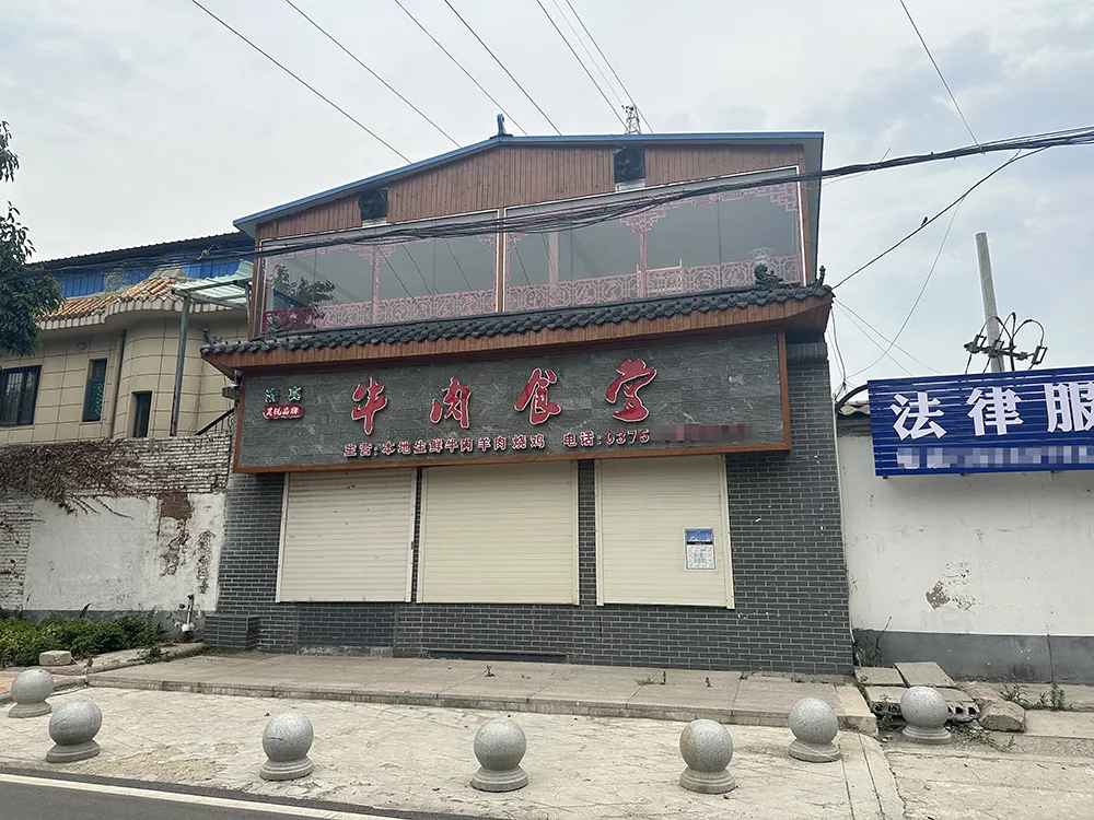一栋两层小楼，招牌写着“牛肉食堂”，卷帘门紧闭，门前摆着石球路障，旁边是一块“法律服务”蓝色招牌。