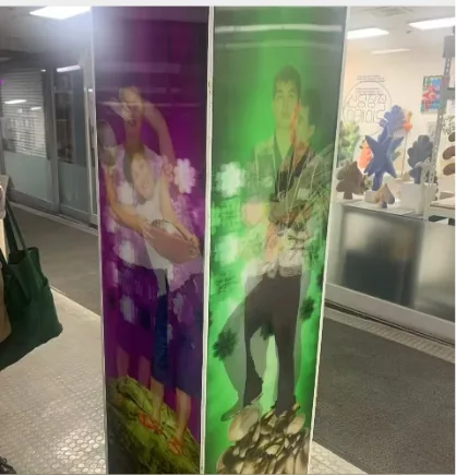 两块紫色和绿色调的透镜印刷展板立在瓷砖地面的通道中，紫色一面印着怀抱婴儿的女性，绿色一面印着站立的年轻男子，背景衬着绿叶图案，通道尽头摆着毛绒玩偶等小物件。
