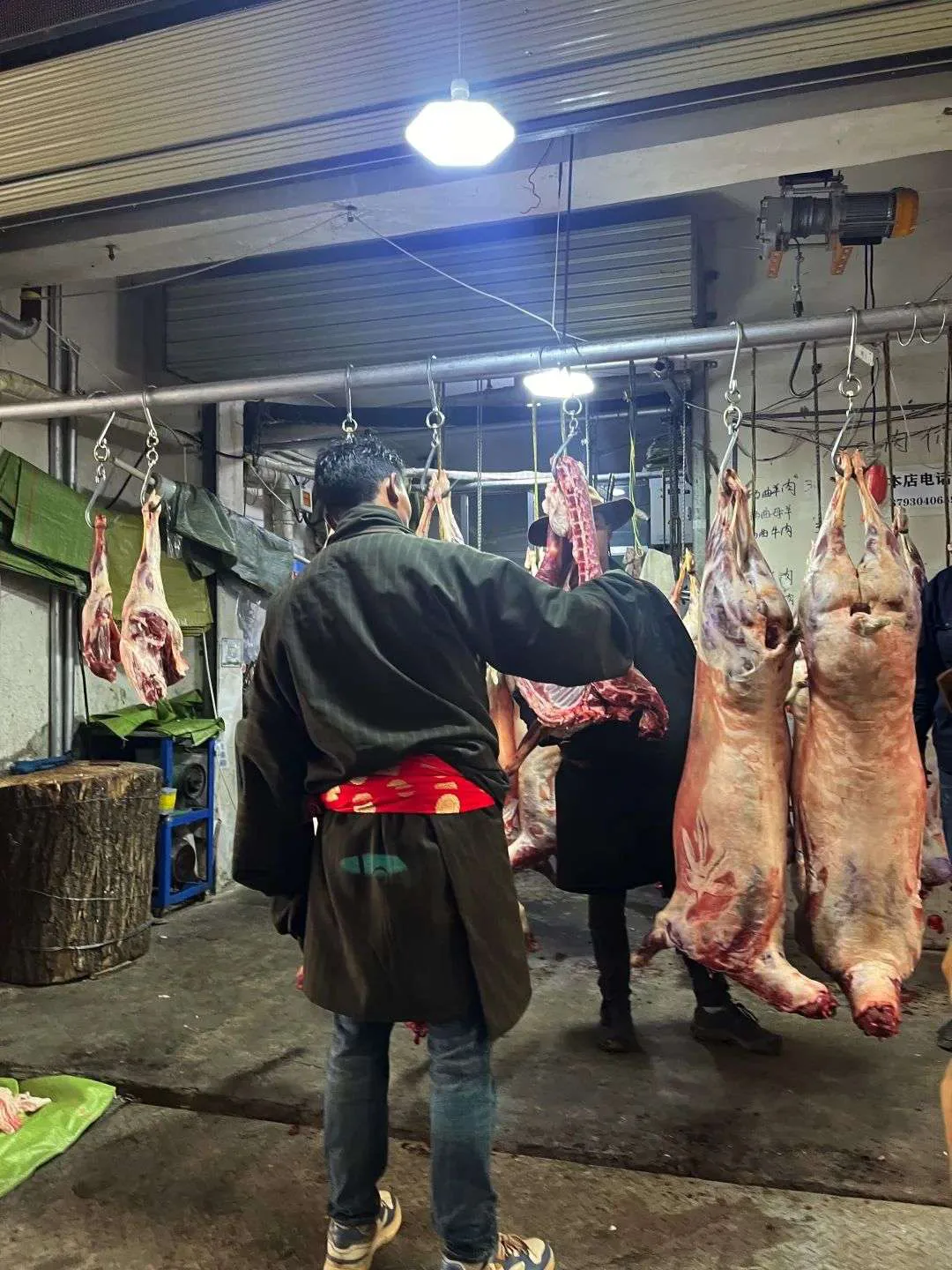 肉铺内几只整只羊胴体悬挂在铁钩上,一位穿藏式外衣的男子背对镜头站在肉架前挑选。